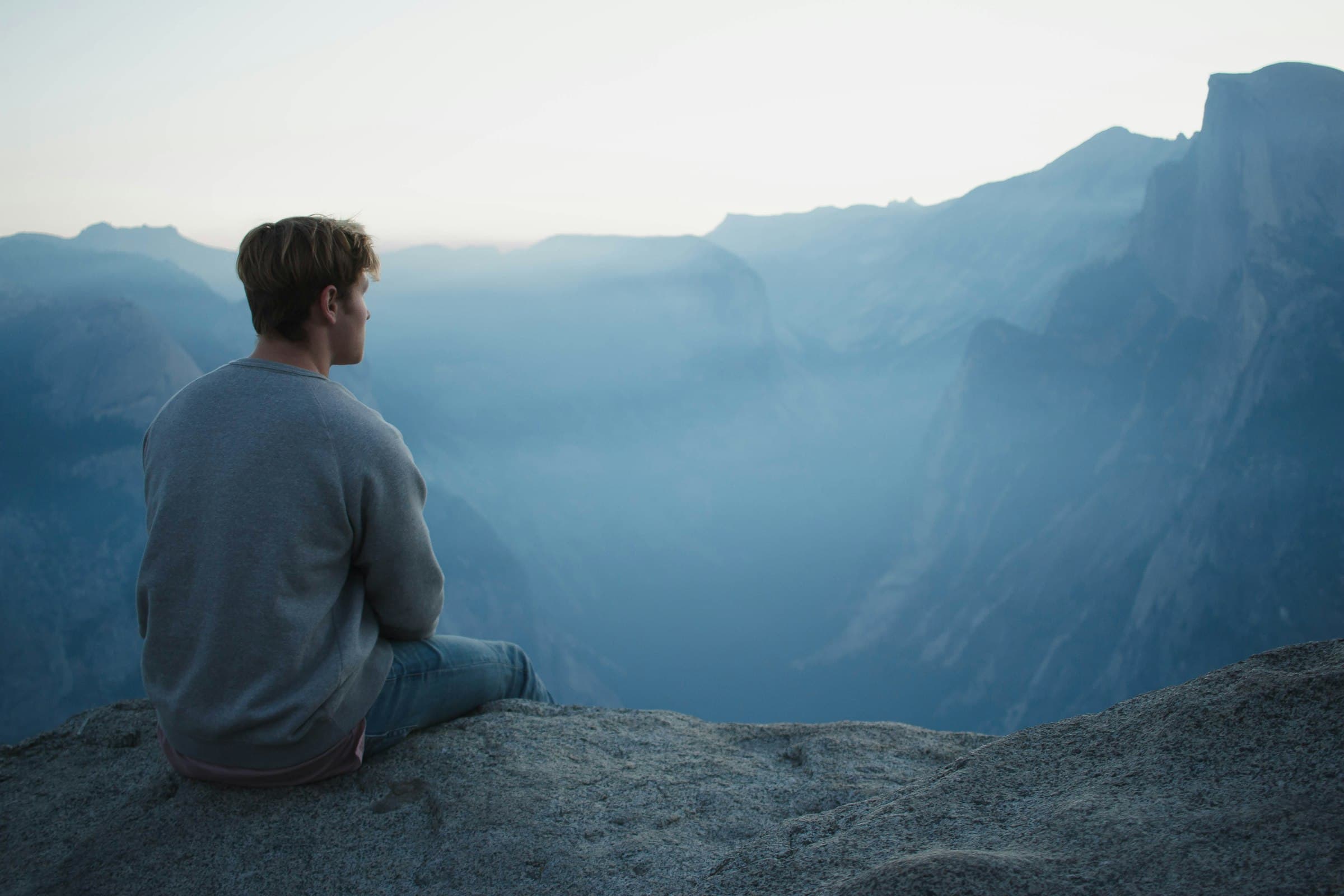 A man sitting on a cliff edge, looking out over misty mountains at dawn