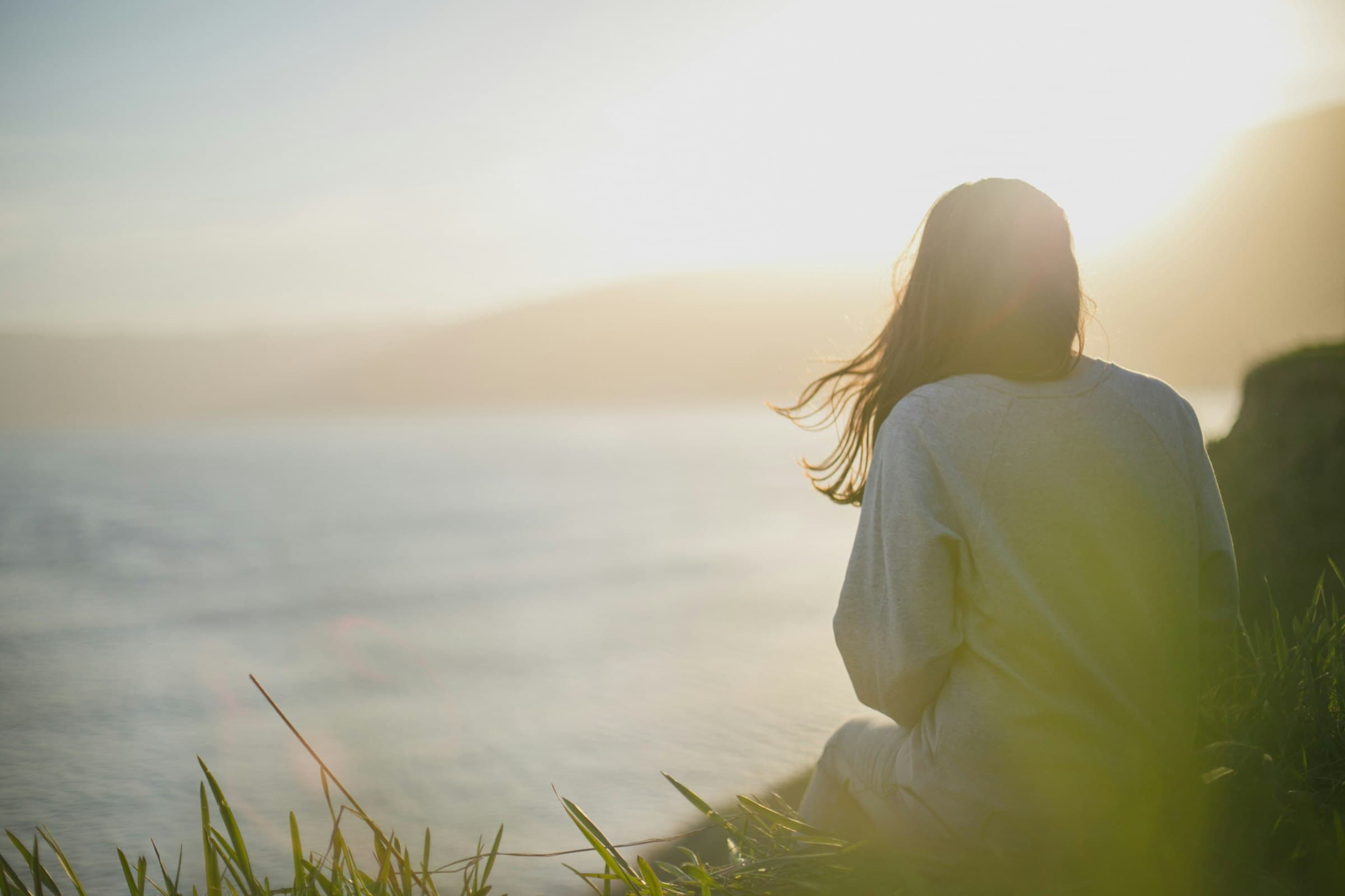 A woman sitting on a grassy cliff, looking out over the sea in soft morning light